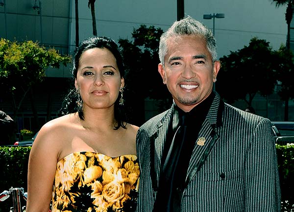 Cesar Millan in a black lined coat with his wife in a black bodycon with yellow flowers printed on it.